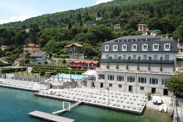 Budova hotelu u jezera obklopená zelení a dalšími domy.