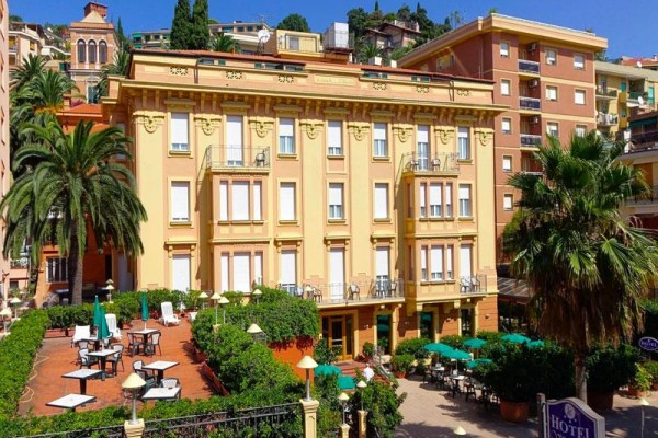Žlutý hotel s terasou a palmami na pozadí města.