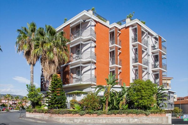 Moderní čtyřpatrová budova s balkóny obklopená palmami a zelení.