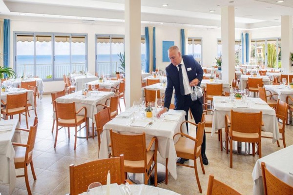 Číšník v obleku upravuje stůl v světlé restauraci s výhledem na moře.