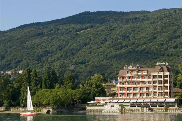 Budova hotelu u jezera s plachetnicí na vodě a zalesněným kopcem v pozadí.