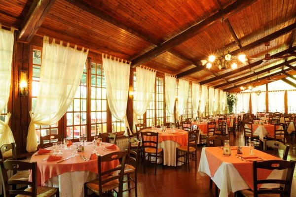 Restaurace s dřevěným stropem, odkud visí lustry, s prostřenými stoly a bílými závěsy na oknech.