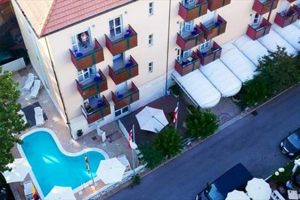 Pohled shora na budovu s bazénem a balkony vedle úzké silnice se zaparkovanými auty. 