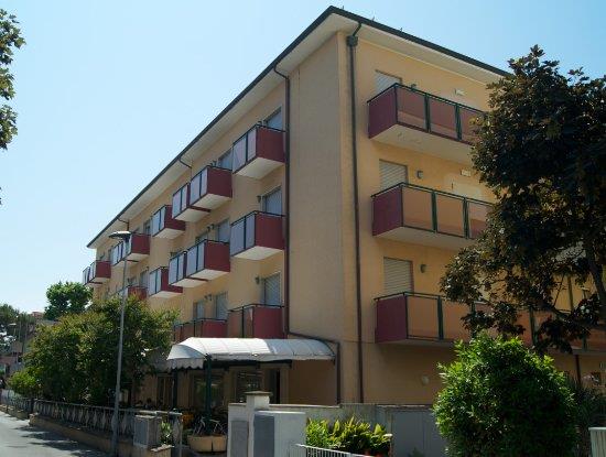 Čtyřpatrový bytový dům s balkony a okolními stromy.  