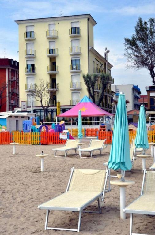 Lehátka a slunečníky na písečné pláži před vícepatrovou budovou a oploceným dětským hřištěm.