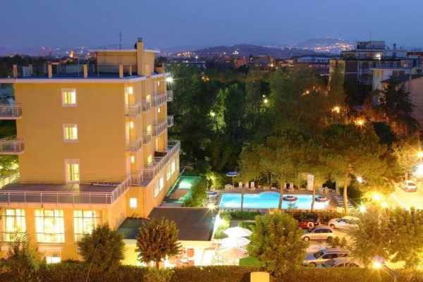 Večerní pohled na osvětlený hotel s bazénem obklopený stromy a městskou krajinou v pozadí.  