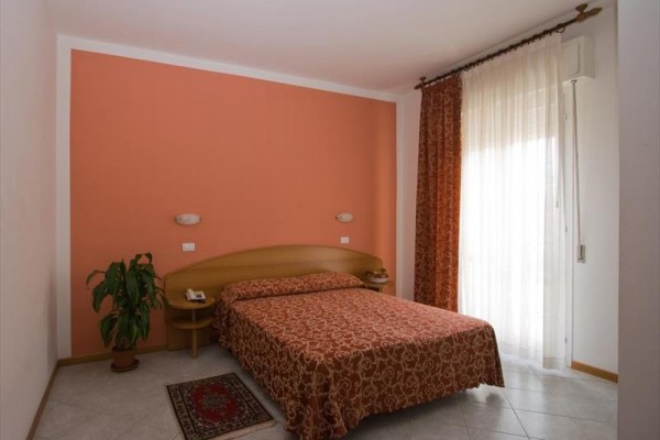 Ložnice s oranžovou stěnou, manželskou postelí, rostlinou a závěsem na okně.  