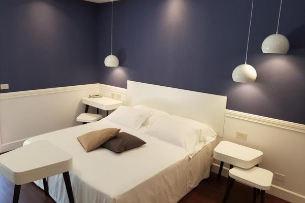 Moderní ložnice s modrou stěnou, bílou postelí a závěsnými lampami.