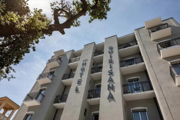 Šedá budova hotelu Quisisana s balkony a stromem v popředí.