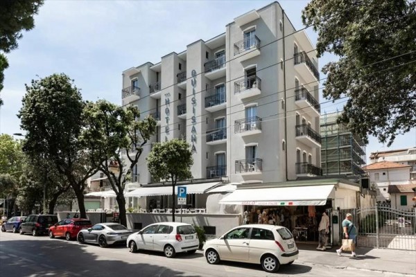 Pětipatrový hotel na rohu ulice s auty zaparkovanými vpředu.