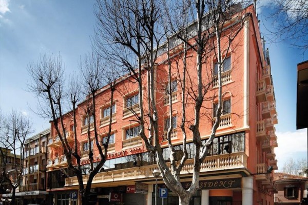 Čtyřpatrová budova s růžovou fasádou a balkonky, před kterou parkují auta a roste několik holých stromů.
