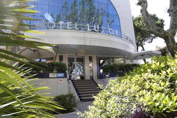 Vchod do hotelu Luxor s moderní skleněnou fasádou a schody lemovanými zelení.