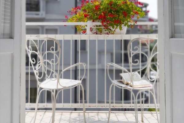 Malý balkon s bílými kovovými židlemi a květináčem s červenými květy.