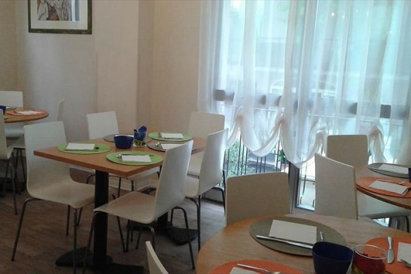 Restaurace s kulatými stoly, bílými židlemi a jasným denním světlem proudícím skrz závěsy.