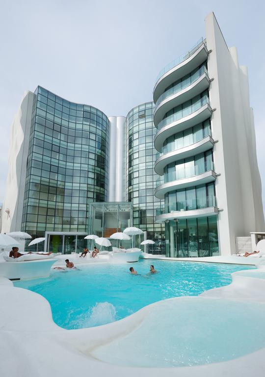 Moderní hotel s bazénem, kolem kterého jsou rozmístěná lehátka a slunečníky.