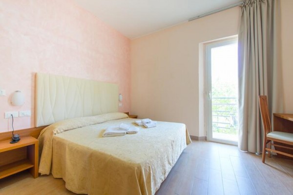 Hotelový pokoj s manželskou postelí a otevřenými dveřmi na balkon.