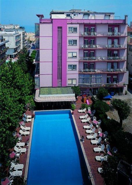 Růžový hotel se čtyřmi patry a venkovním bazénem obklopeným lehátky.
