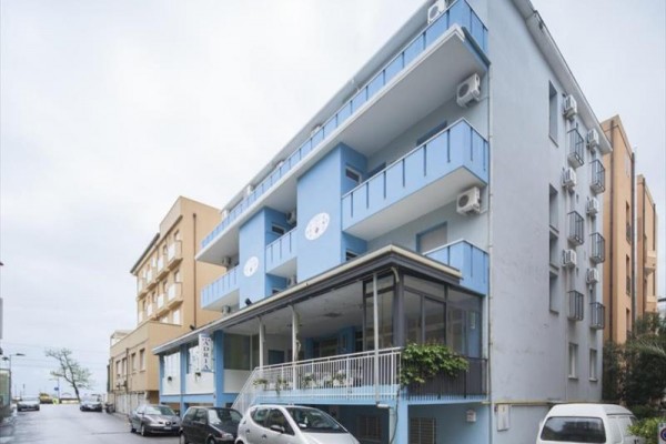 Moderní modro-bílá třípatrová budova s balkóny a zaparkovanými auty před vchodem.