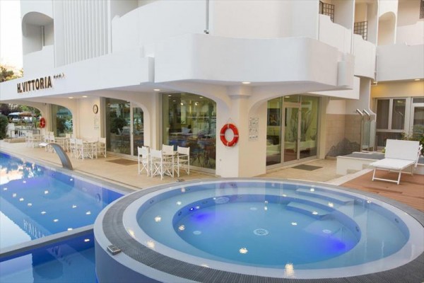 Venkovní bazén s vířivkou u hotelu s terasou vybavenou bílými židlemi a stolky.
