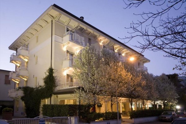 Detail osvětleného hotelu s balkonky a kvetoucími stromy v popředí za soumraku.