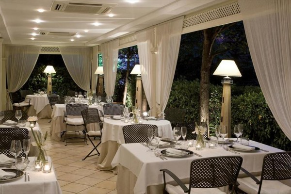 Elegantní restaurace s bílými ubrusy, elegantními židlemi a stojacími lampami u oken s výhledem do zahrady.