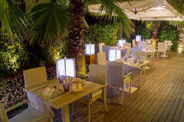 Venkovní restaurace s prostřenými stoly, obklopená palmami a osvětlená lampami.