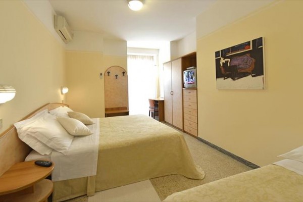 Jednoduše zařízený hotelový pokoj s manželskou postelí, další postelí, televizí a obrazem na stěně.  