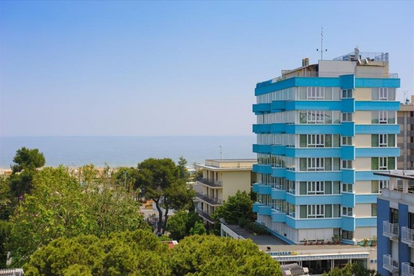 Modro-bílý vícepatrový hotel u moře, obklopený zelení.
