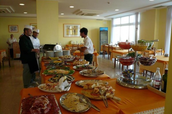 Hotelová restaurace s bufetovým stolem plným různých pokrmů a kuchaři v pozadí.