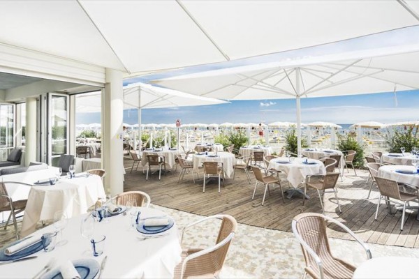 Restaurace s venkovní terasou, zakrytou bílými slunečníky, s výhledem na pláž.