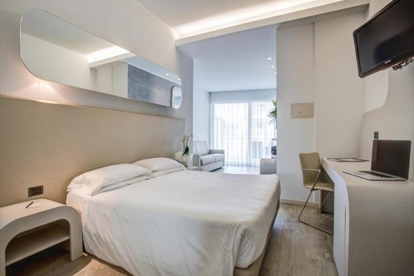 Moderní hotelový pokoj s manželskou postelí, psacím stolem, křeslem a balkonem.