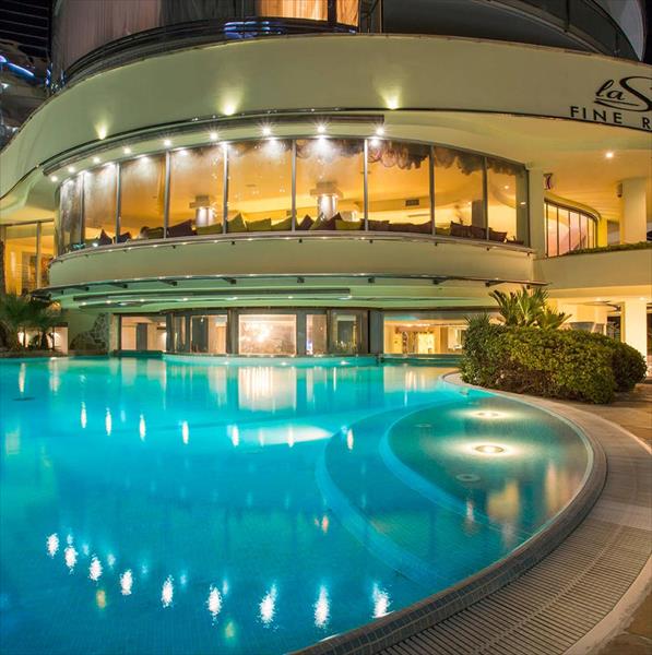 Noční osvětlený bazén u moderního hotelu s kulatou skleněnou restaurací.