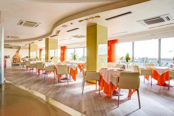 Interiér světlé moderní restaurace s oranžovými ubrusy a velkými okny.