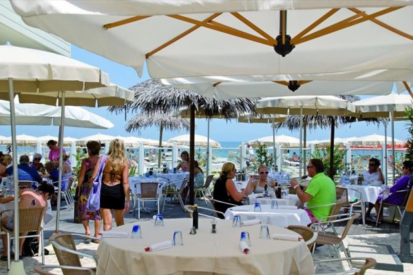 Lidé sedí u stolů pod slunečníky v plážové restauraci s výhledem na moře.  