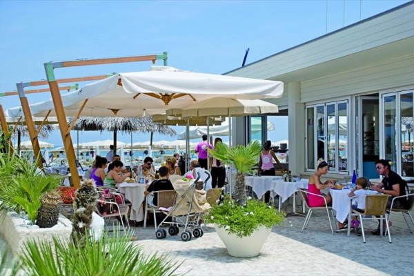 Lidé sedí u stolů pod slunečníky na venkovní terase restaurace u pláže.