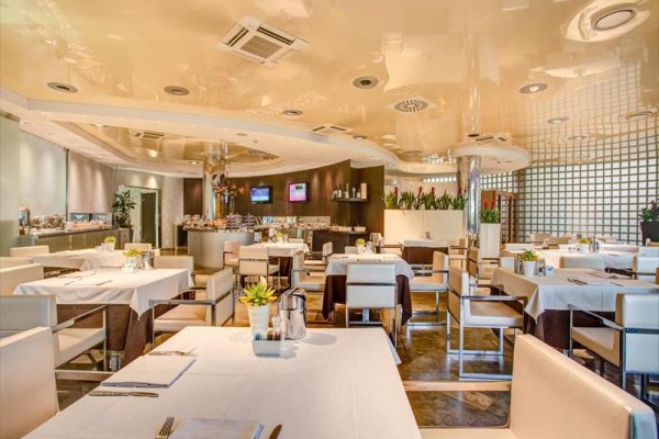 Moderní restaurace s bílými stoly a židlemi, výzdobou rostlin a televizory na stěně.