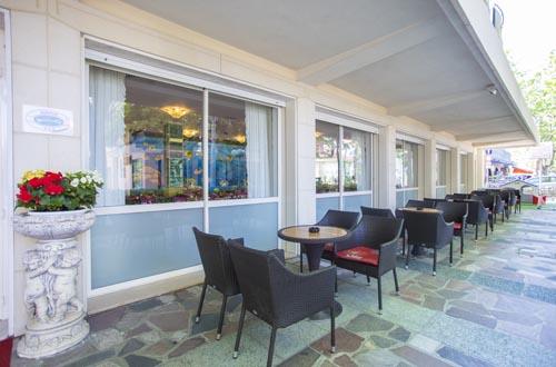 Venkovní posezení restaurace s černými židlemi a kulatými stoly u velkých oken.