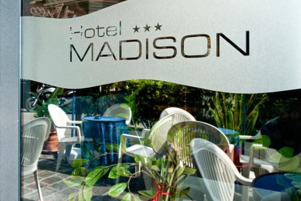 Logo hotelu Madison na skleněných dveřích s odrazem stoliček a rostlin.