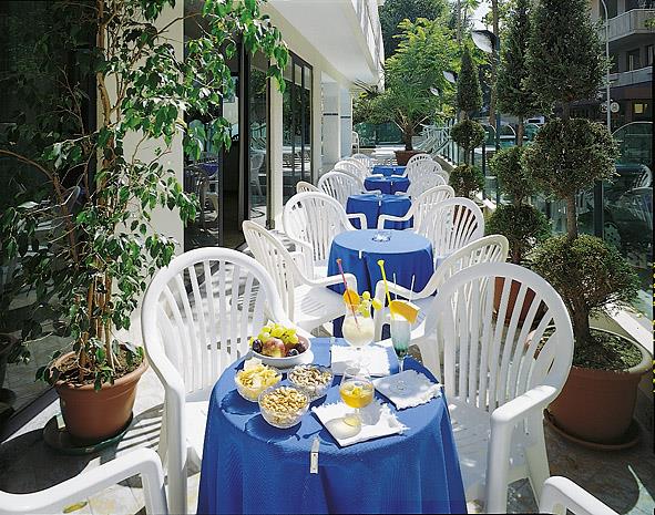Venku restaurace s modrými ubrusy, bílými židlemi a jídlem na stolech.