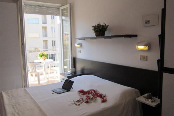 Ložnice s manželskou postelí, otevřenými dveřmi na balkon a stůl s kytkou v květináči venku.
