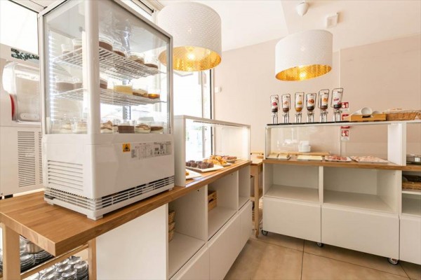 Interiér samoobslužného bufetu s vitrínami na zákusky a dávkovači na cereálie pod moderními lampami.