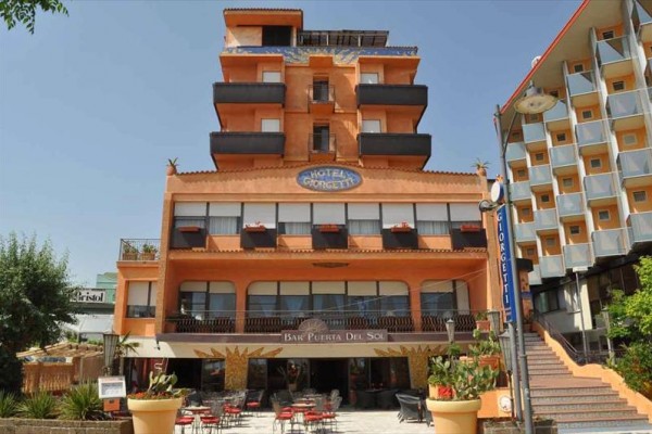 Oranžová budova hotelu s názvem nad vchodem a předzahrádkou s posezením.