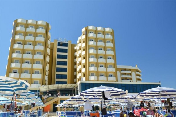 Velký hotel s balkony a modrobílými plážovými slunečníky na písečné pláži.