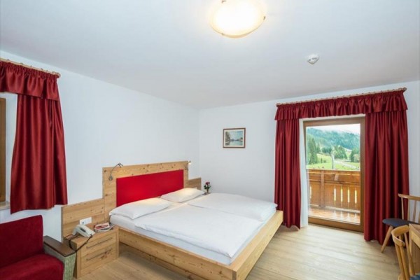 Pokoj s dřevěnou postelí, červenými závěsy a balkonem s výhledem na krajinu.
