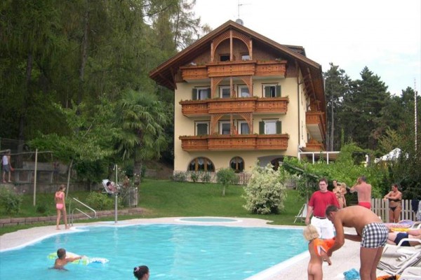 Dřevěná horská chata s balkonem u bazénu, kolem něj lidé relaxují.