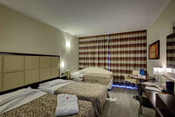 Hotelový pokoj se dvěma postelemi, stolem a závěsy na oknech.