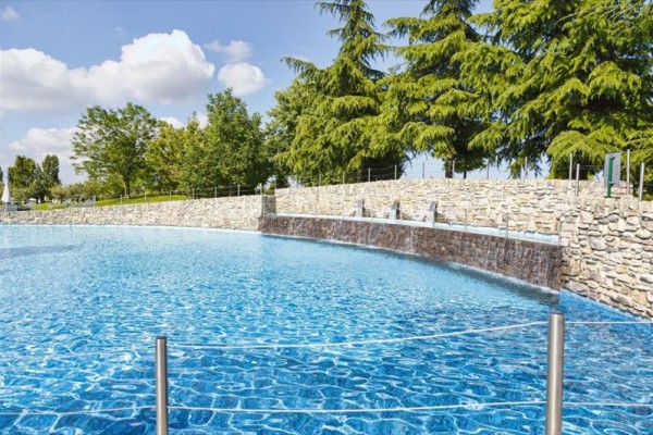 Modrý bazén s vodopády obklopený kamennou zdí a stromy v pozadí.