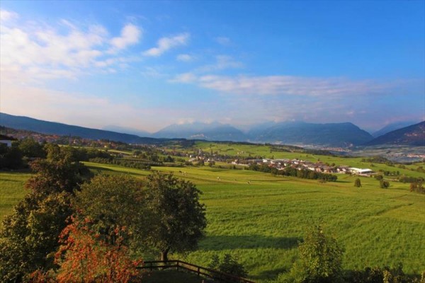 Rozlehlá krajina s loukami, stromy a horami v pozadí.