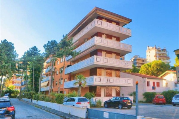 Čtyřpodlažní obytná budova s balkóny obklopena stromy a auty na ulici.