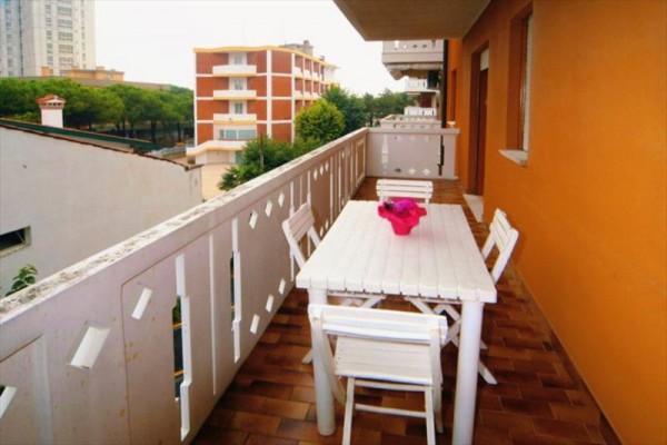 Dlouhý balkon s plastovým stolem, čtyřmi židlemi a květináčem s růžovými květinami, výhled na stromy a budovy.